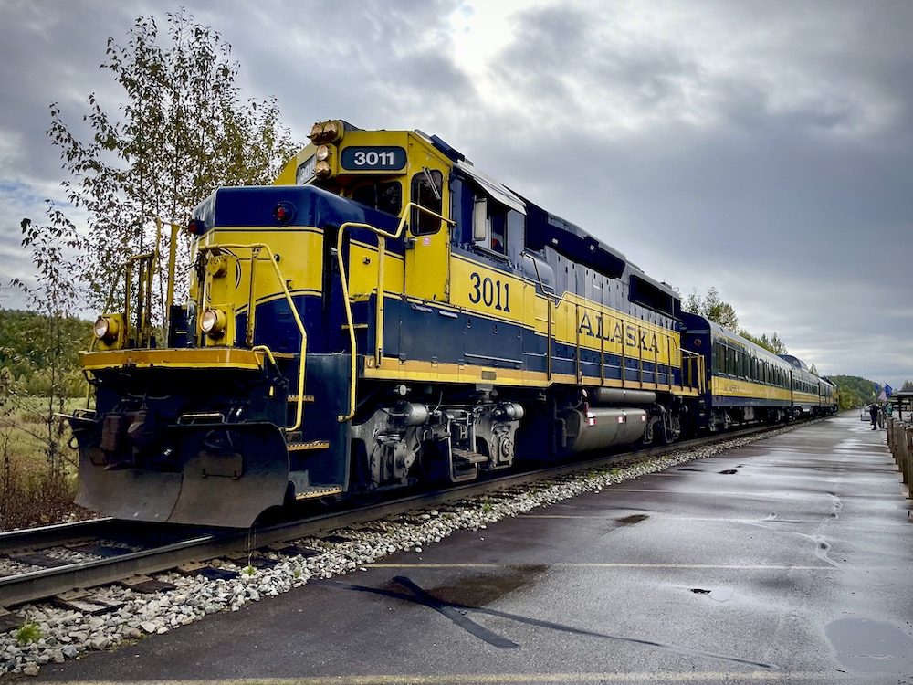 A blue with yellow stripe diesel locomotive and short train next to a rain-soaked platform. Alaska by rail tour.