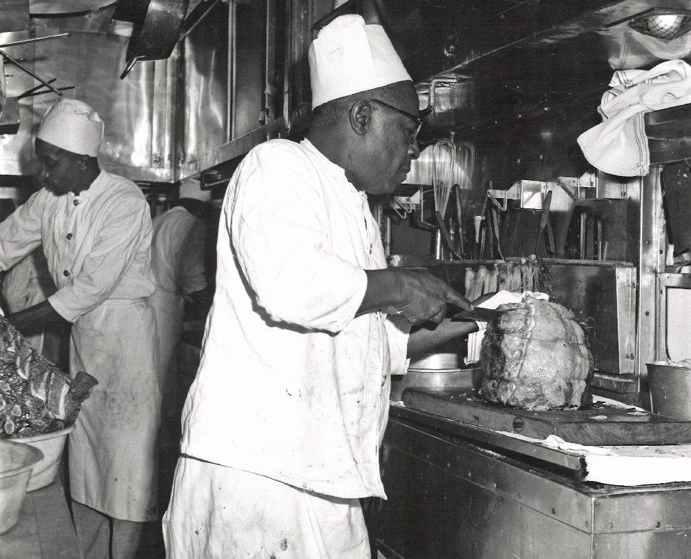Chef carving prime rib in dining car kitchen.