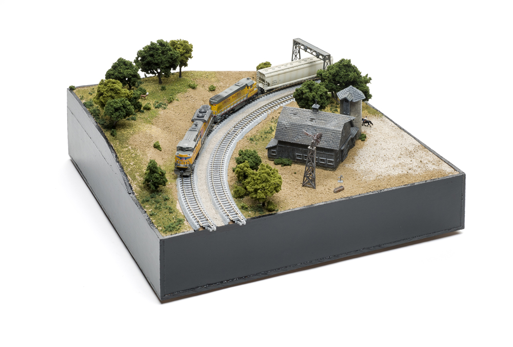Small model train scene with two curving gray tracks, a weather-worn wood barn, a few green trees, and two yellow locomotives.