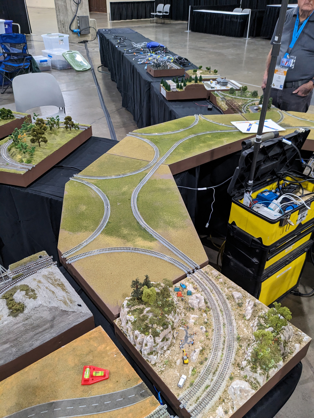 Green and brown boxes of different shapes with small gray railroad tracks on top set up on tables with black drapes over them.