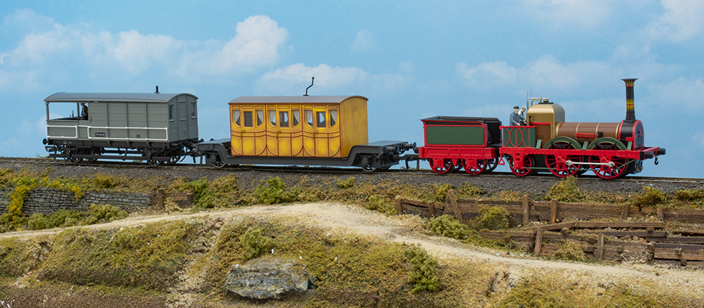 Rapido UK Titfield Thunderbolt: Small, colorful model train on scenery base with green grass, brown road, and bare wood cribbing.