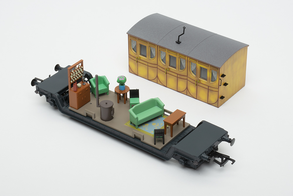 Black model flat car with yellow railway coach body removed showing small living room with couch, table, chairs, and China cabinet with white dishes.