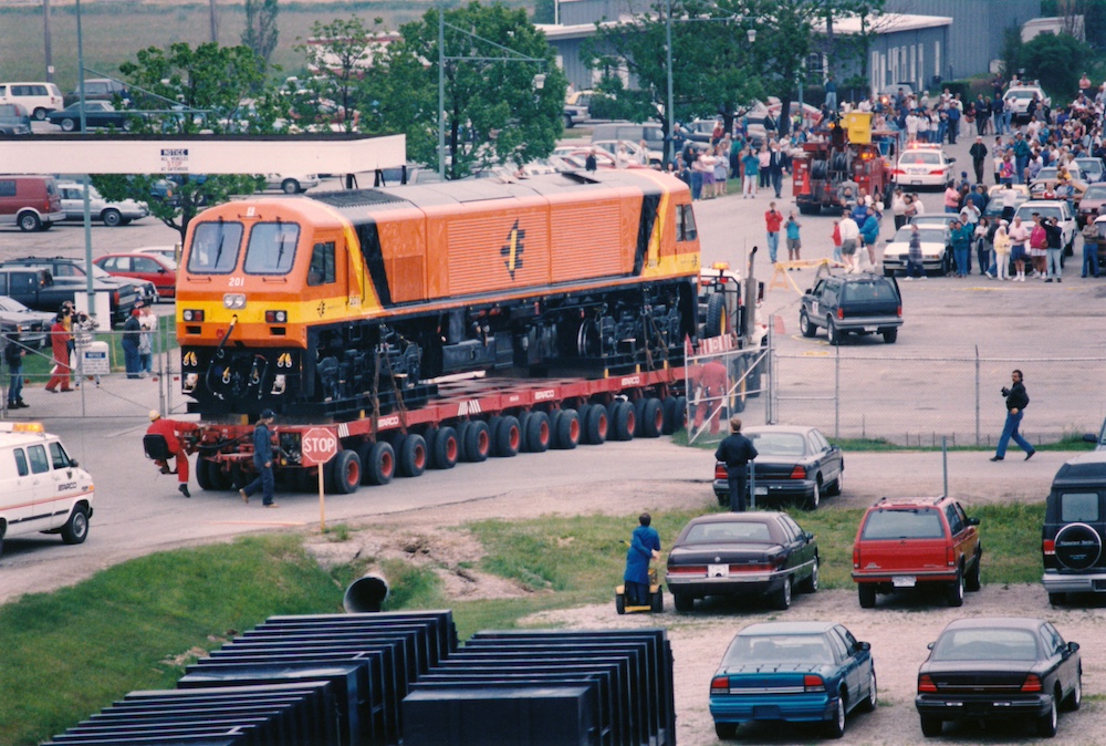 An orange European-design diesel locomotive moves down an urban street on a multi-wheeled trailer, witnessed by many people watching from the sidewalks