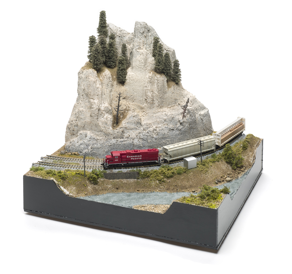 Small black box holding model scene with tan mountain, green trees, and a red train.