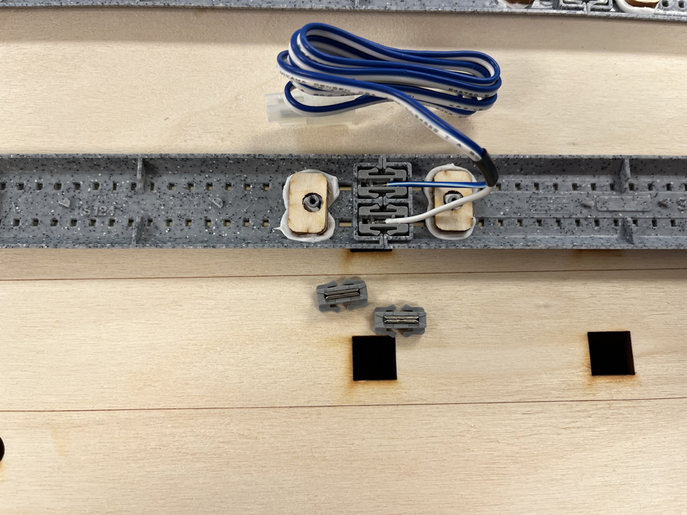 Small gray pieces of plastic and one long section of gray plastic with blue and white wires attached to it resting on a bare wood surface.