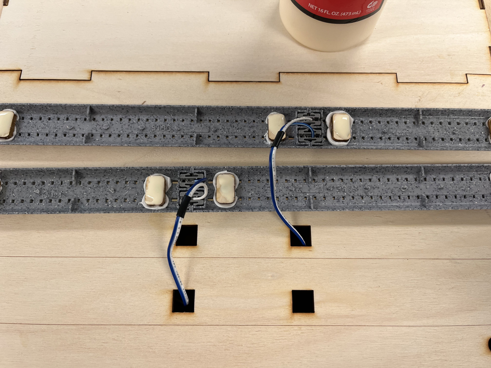 Two gray strips of plastic model train track with blue and white wires attached resting on a bare wood surface. The strips have blobs of wet yellow glue on them, and the wires pass through square holes in the wood surface.