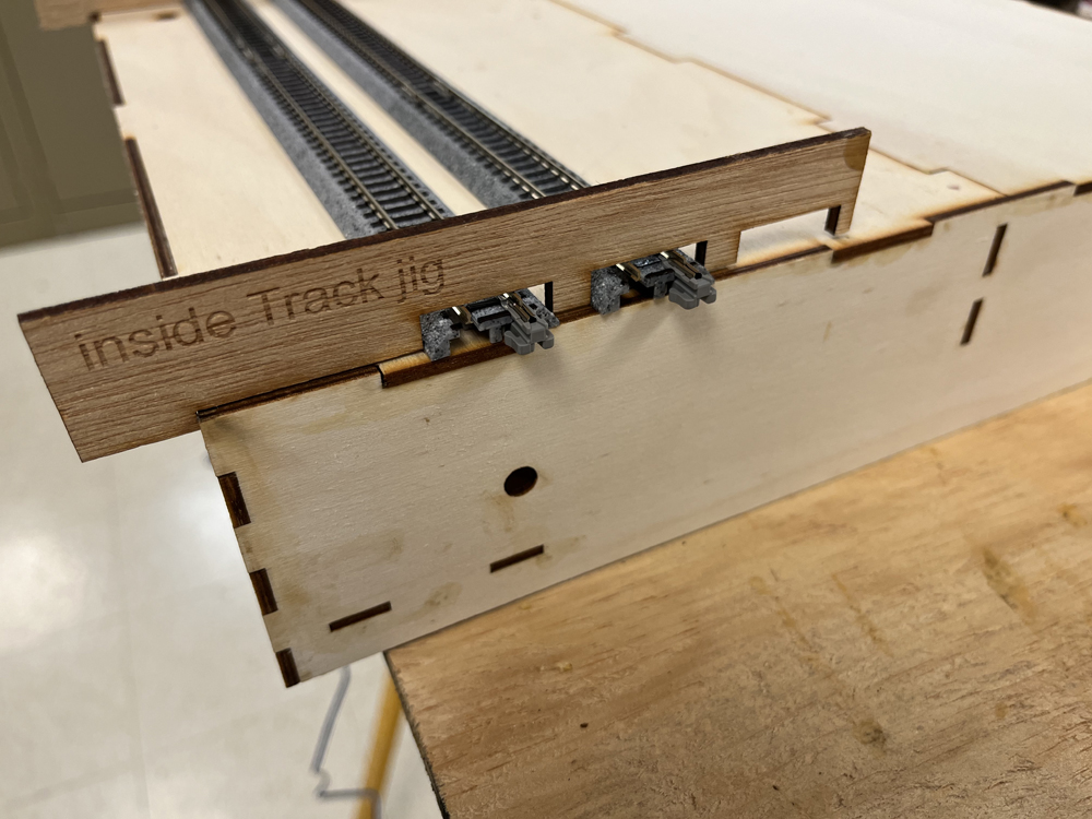 Two gray strips of plastic model train track with notched wood spacer between them keeping them at a uniform distance.