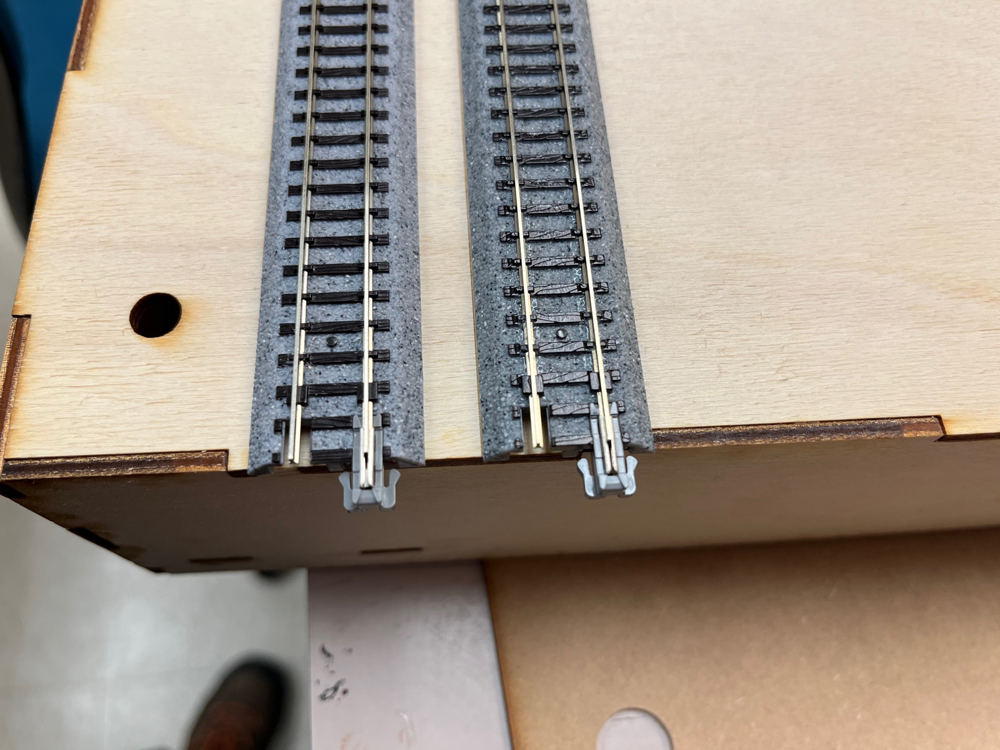 Two gray strips of plastic model train track with silver rails and black crossties on a bare wood surface.