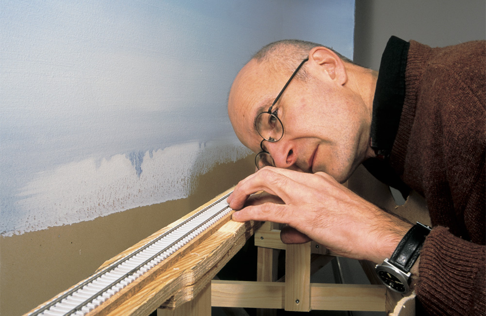 A man lowers his head to track level to check a model railroad track for straightness