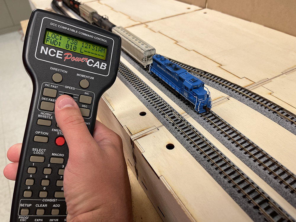 Hand holding black and gray remote control with green lighted screen with blue and gray model train on silver, gray, and black model train track.