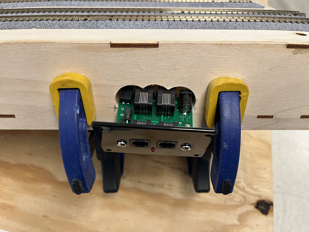 brown wood panel with round holes cut into it to fit green and black circuit board. Also shown are two blue and yellow clamps attached to the wood panel.