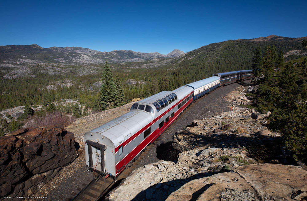 Two private cars behind an Amtrak train in the mountains.
