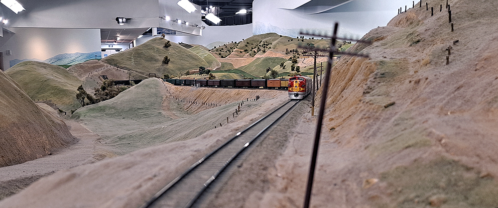 A freight train led by Santa Fe EMD F units in Warbonnet paint climbs a single track through high desert scenery on a large model railroad.