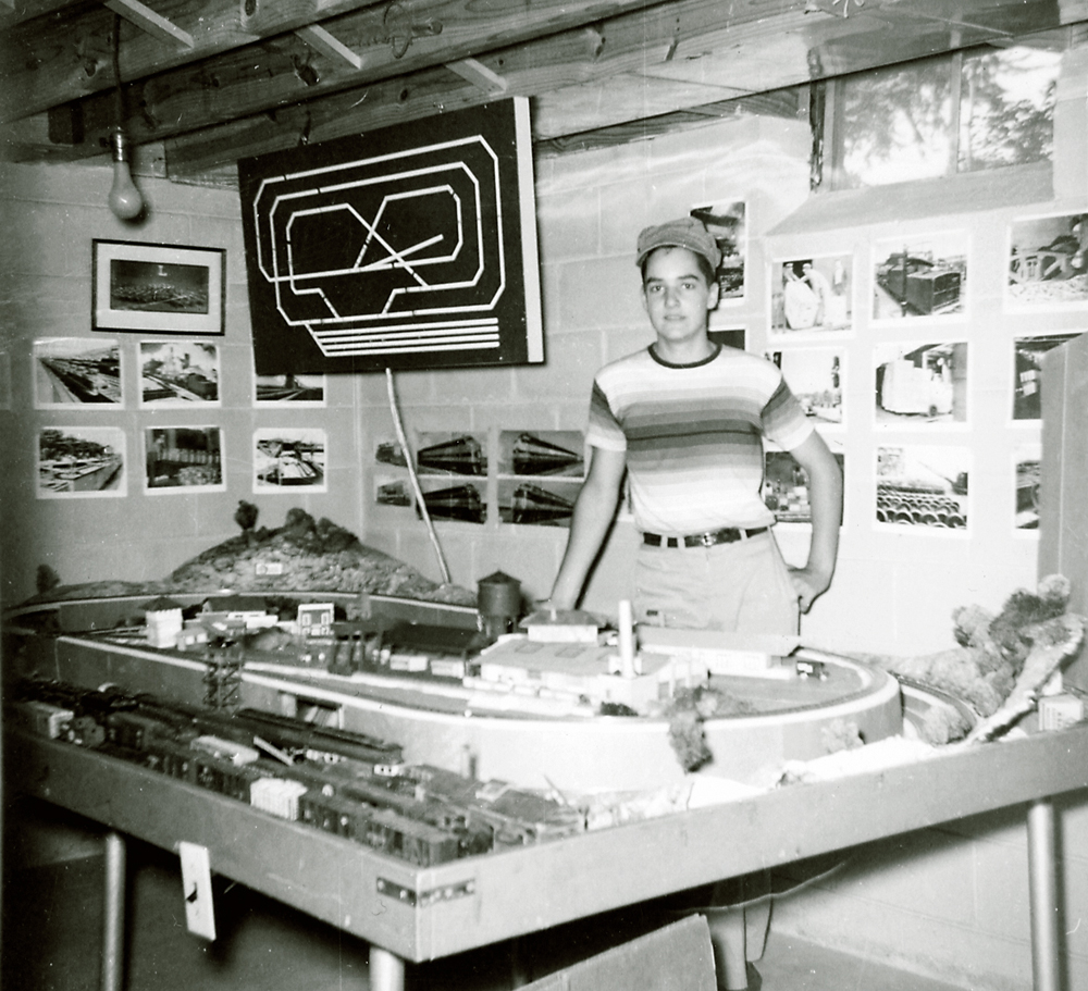 Black-and-white photo of teenage boy in engineer's hat by model railroad.