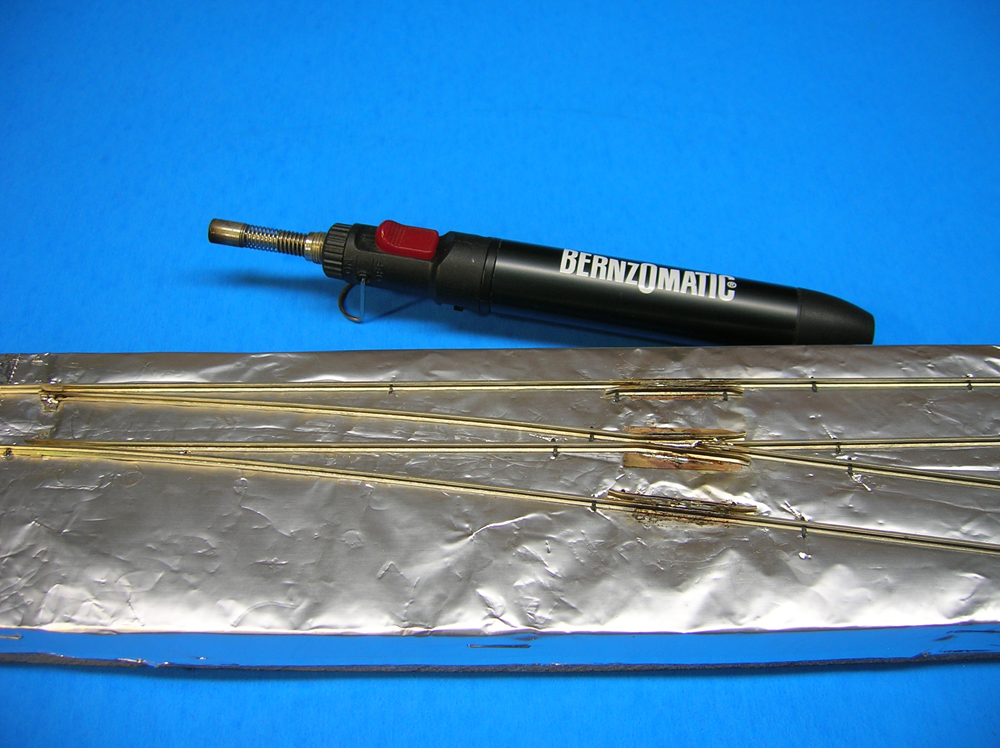 A propane torch next to rails arranged for a turnout on a foil-covered piece of wood