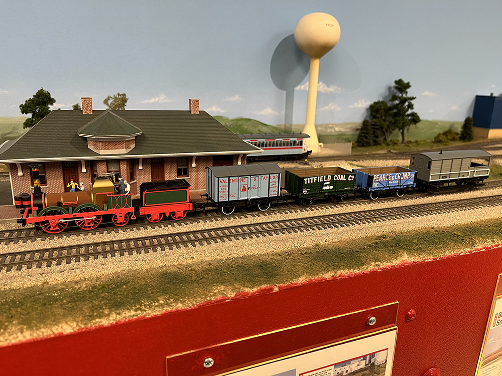 Small model British train with brown, red, and green steam locomotive pulling four small wagons in blue, green and gray.