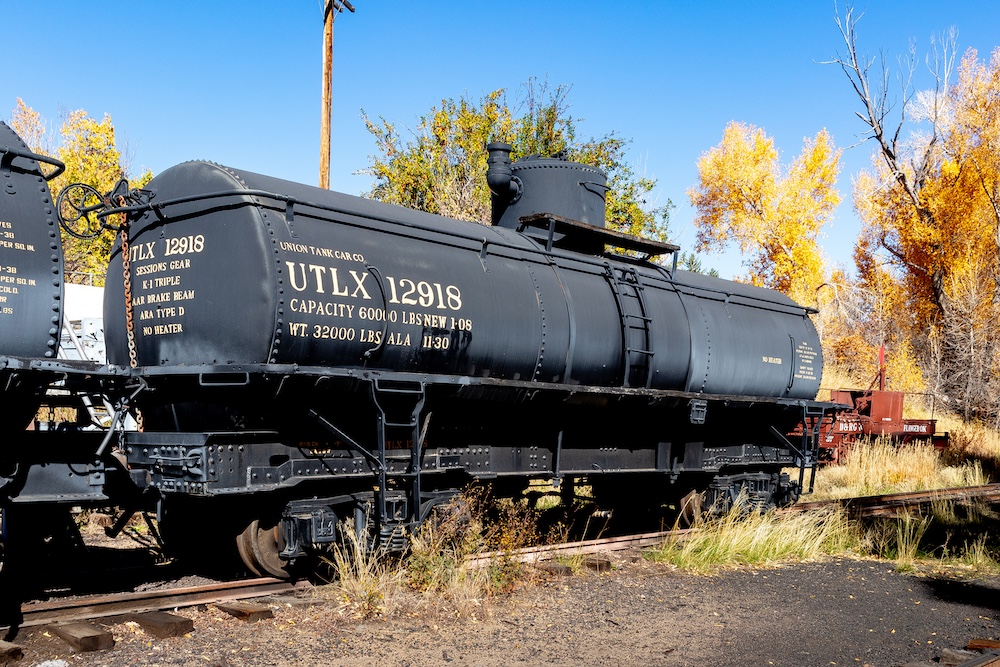 An old black tankcar is parked oa a weed overgrown siding