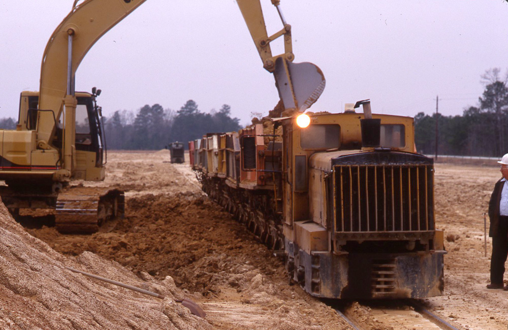 A man watches as a yellow excavator loads clay into hopper cars