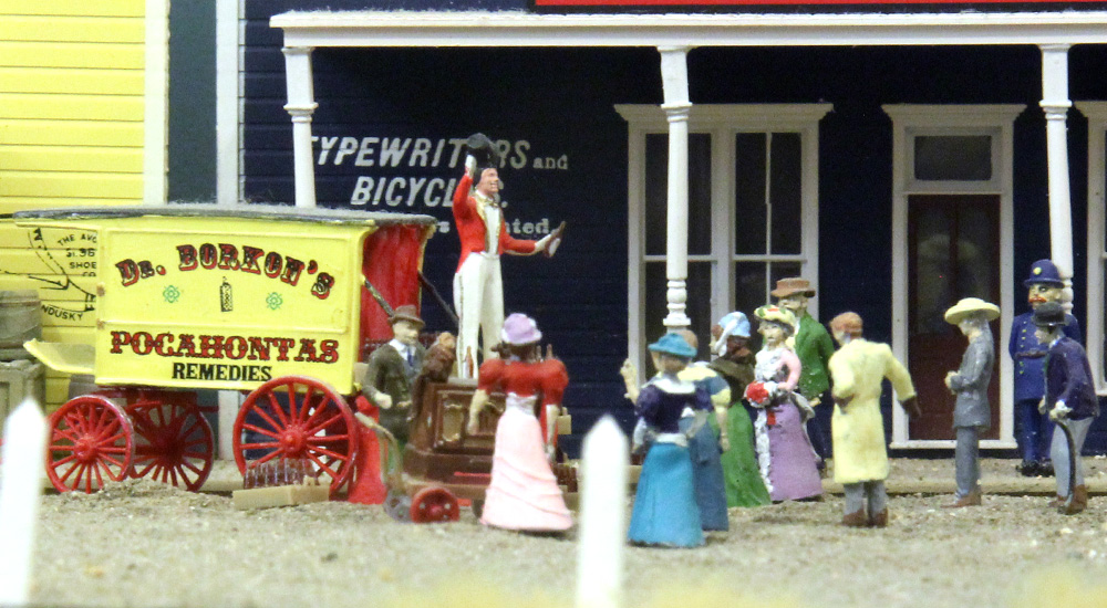A man stands atop a horse-drawn wagon hawking snake oil to an assembled crowd