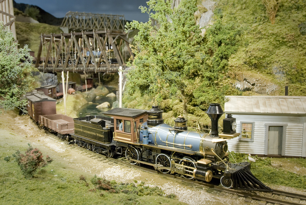 A large scale early-period steam locomotive hauls a long train under a bridge that another train is on.