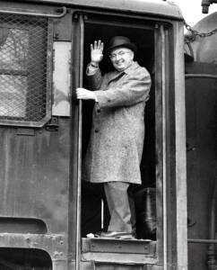Man in winter coat and fedora stands in steam locomotive cab door waving, black and white