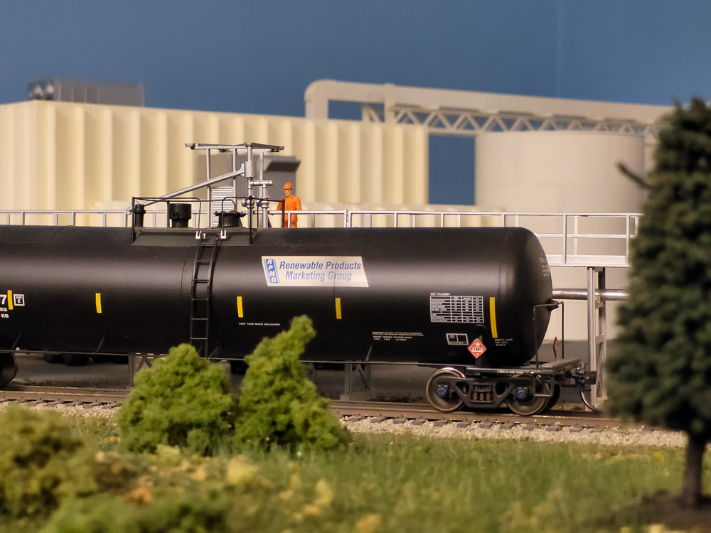 A model figure clad in orange stands next to a black model tank car