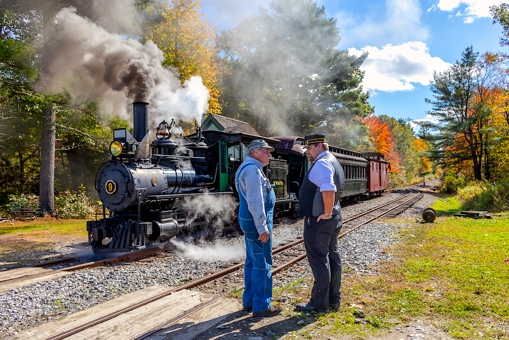 Two men talk next to railroad tracks. A steam locomotive is in the background.