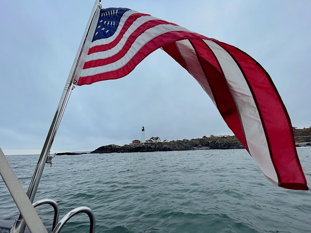 An American flag against a gray sky and water with a lighthouse in the distance