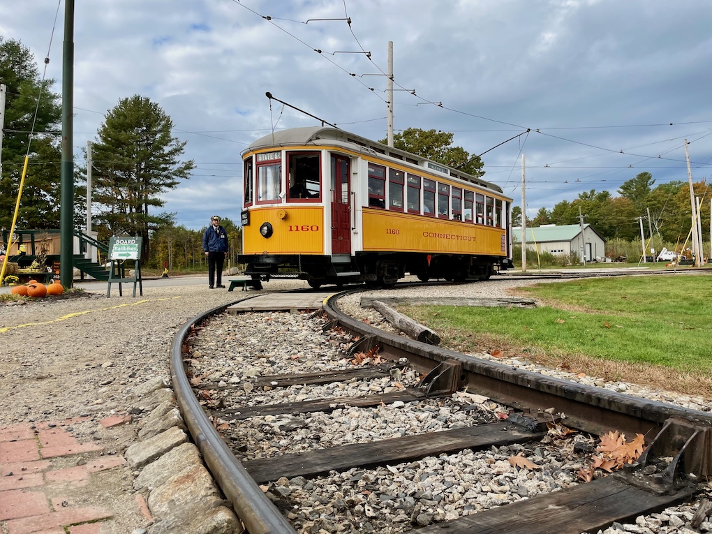 A yellow street car parked on a curving section of track. A man waits next to the car
