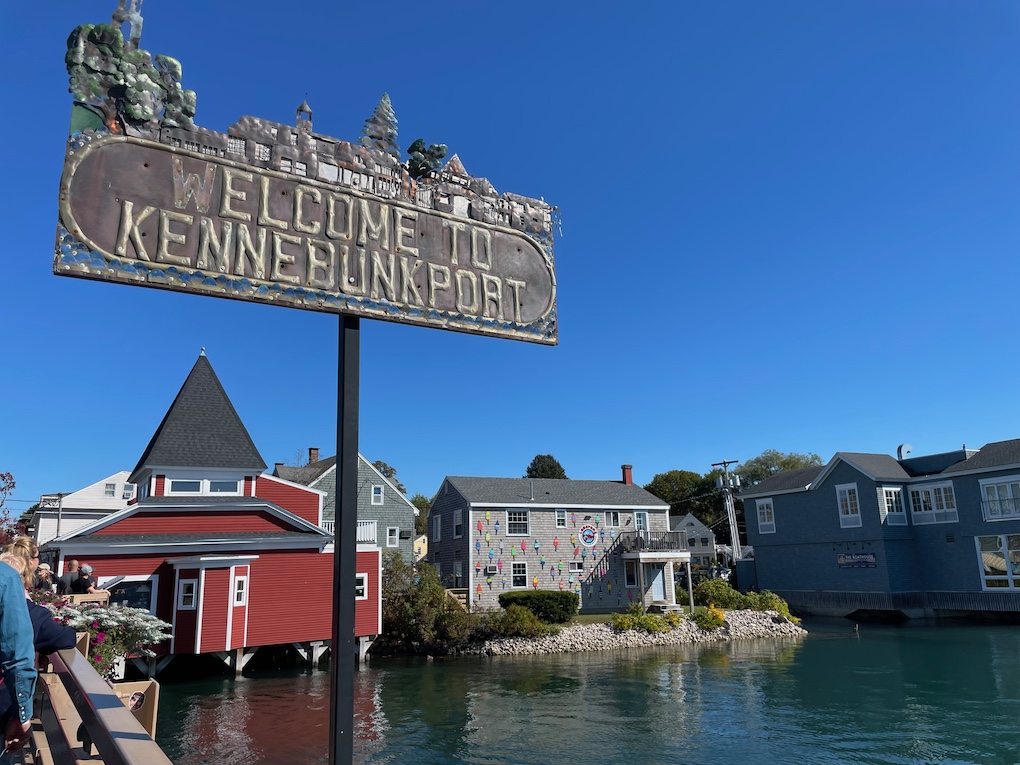A metal sign reading Welcome to Kennebunkport with waterfront buildings behind it