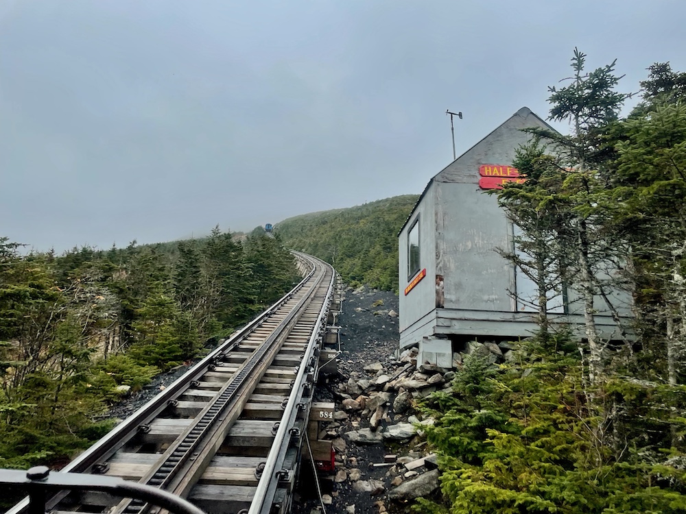 Evergreen trees and a small gray hut frame a steeply climbing railroad