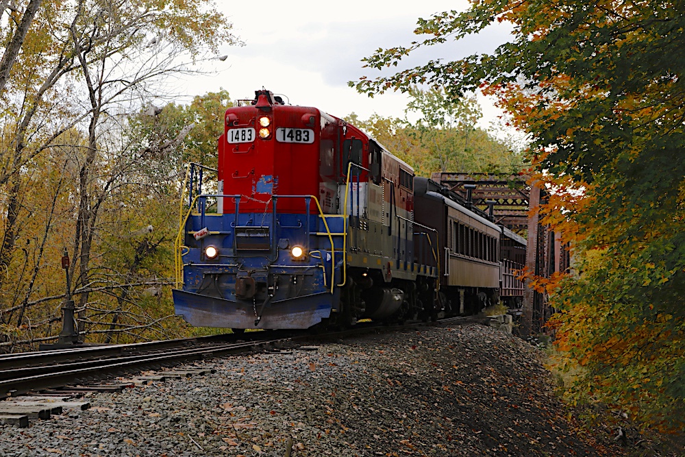 A red, white and blue locomotives crosses a truss bridge with a passenger train in tow