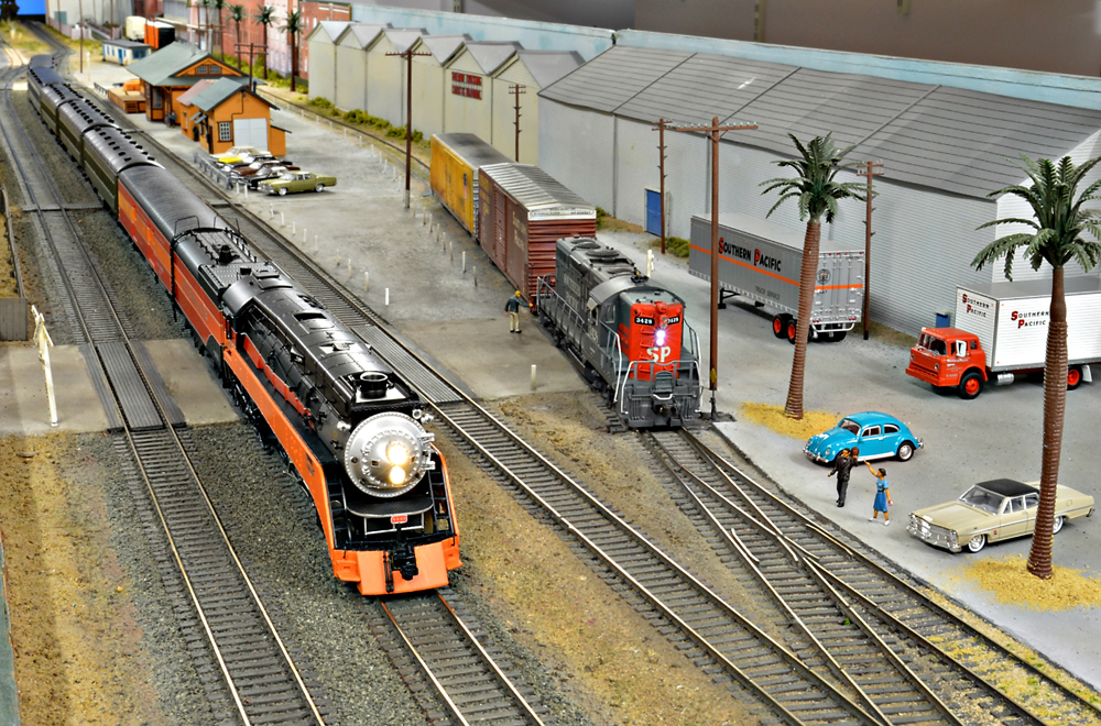 An orange and black model locomotive is seen at left of the image while a more modern locomotive and freight car is seen at center of the image on a model railroad layout