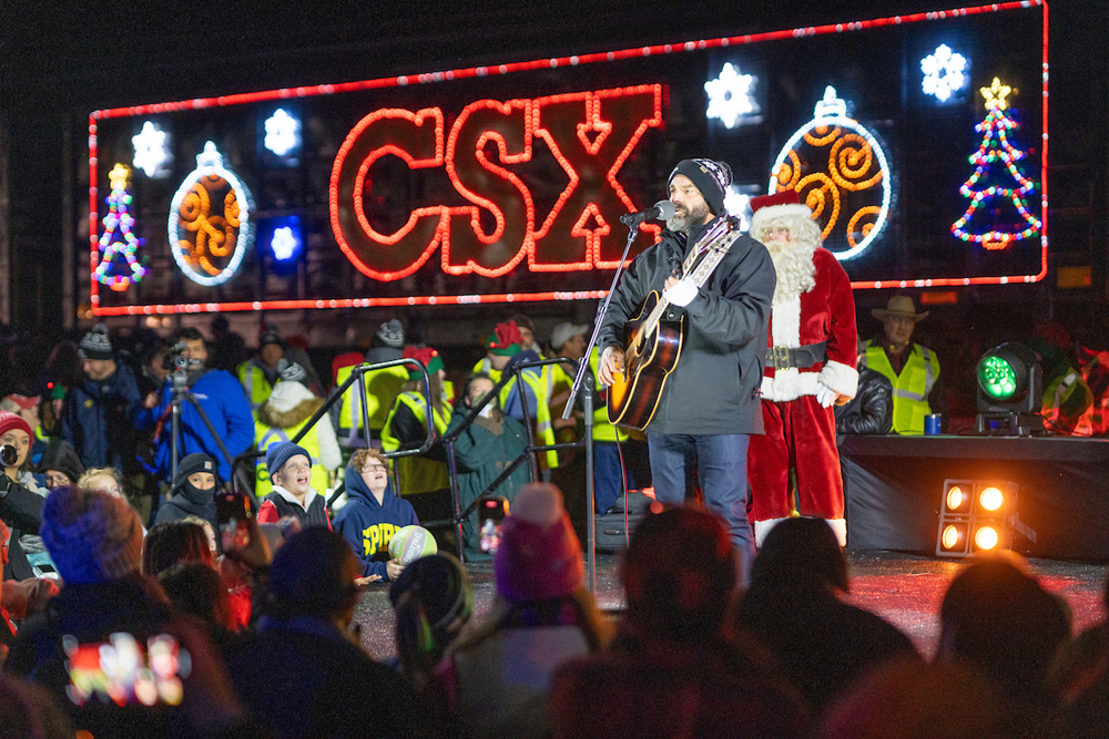 Man with guitar on stage in front of train with holiday lights, with Santa Claus standing in background