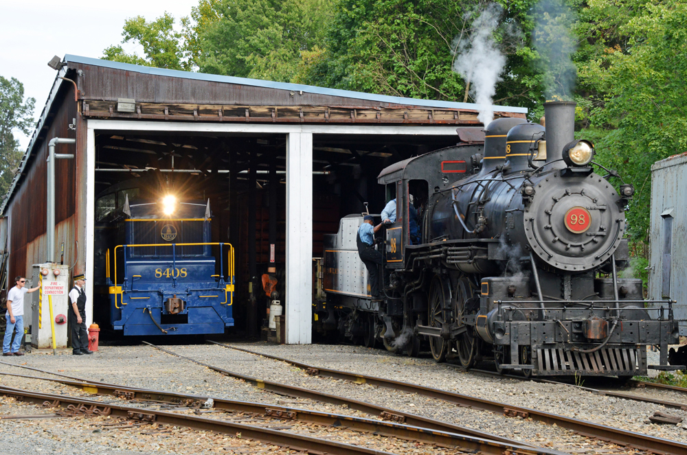 Steam locmotive outside of enginehouse with diesel switcher inside