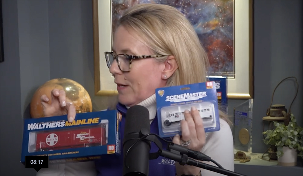 Woman with blond hair and glasses holding two small blue packages containing model train products.