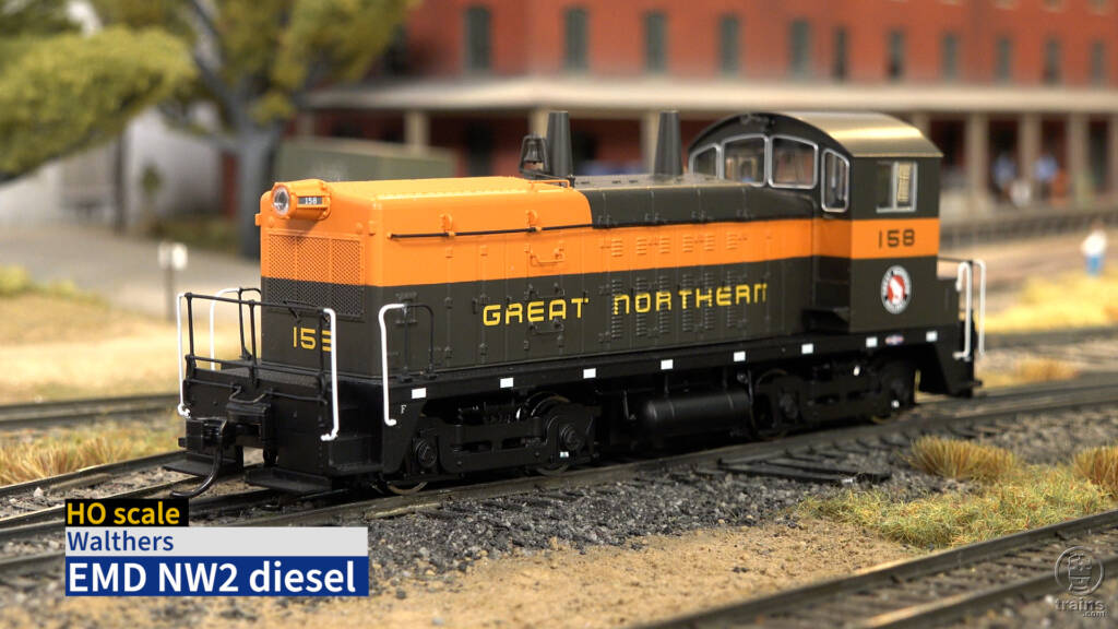 Screen capture from product review video with HO scale end-cab switcher painted orange, green, and black on scenicked layout with title bar in lower left corner.