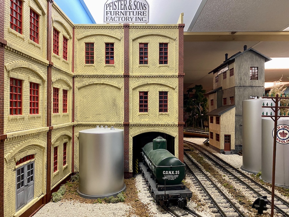 Large-scale green tank car sits near the entrance of a cream scratch-built building