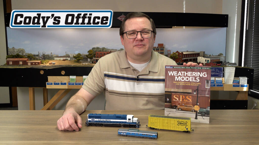 Man in tan shirt with stripes wearing glasses holding book with model trains in foreground