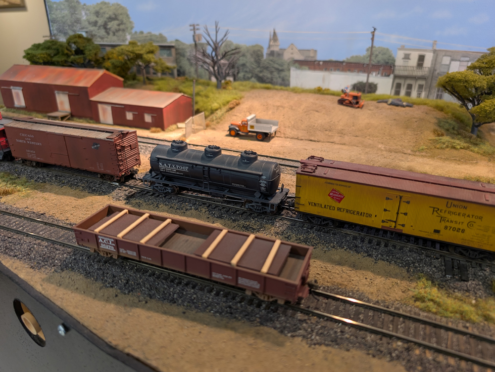 An HO scale reefer, three dome tank car, and boxcar behind a gondola with rebar load