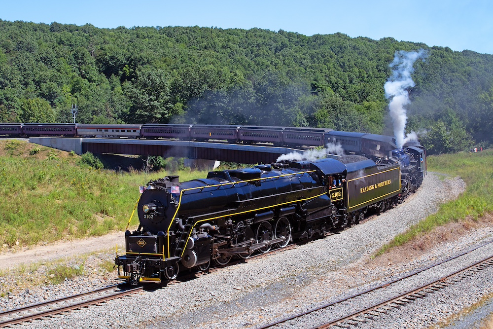 Two steam locomotives cross a bridge and round a tight curve on a summer day