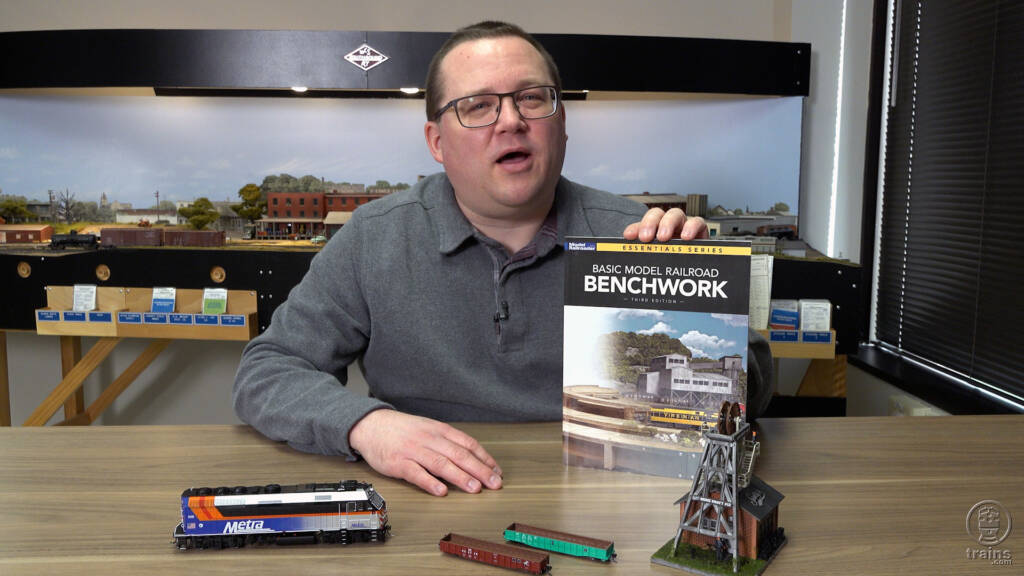 Man in gray shirt wearing glasses holding book with model trains on table.