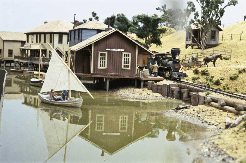A boat sails in a body of water as a train passes on a model railroad layout