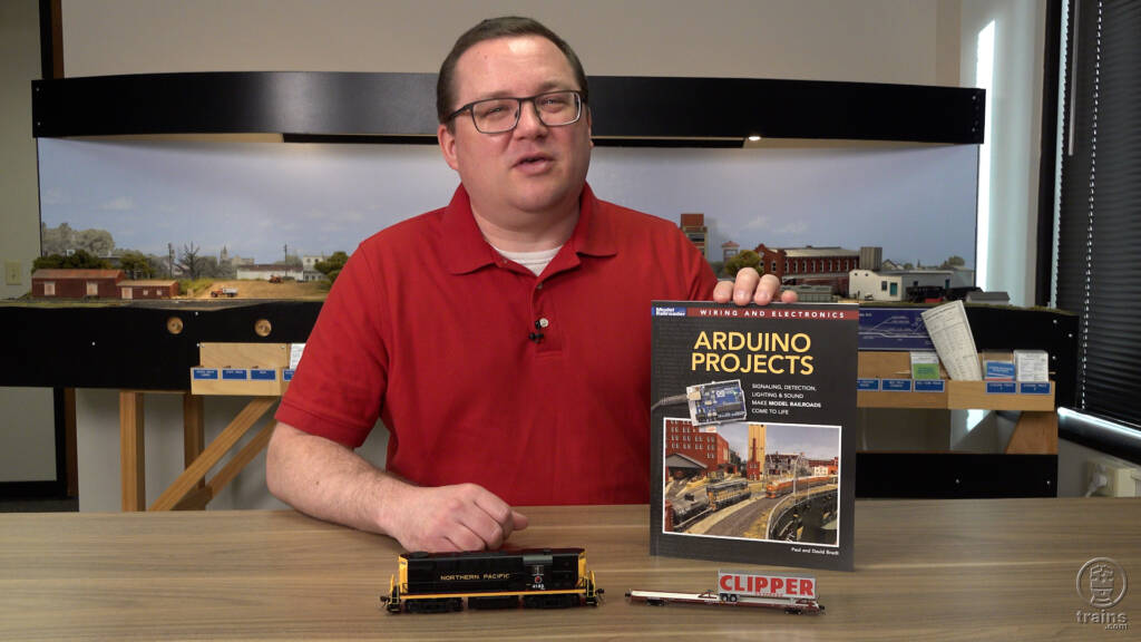 Picture of man in red shirt wearing glasses holding book with model trains in foreground.