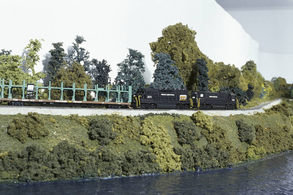 Two black diesel locomotives lead a freight train past a body of water on a model railroad layout