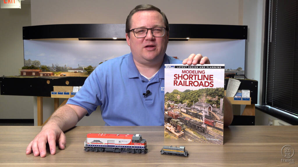 Picture of man wearing blue shirt and glasses holding a book with HO and N scale locomotives in foreground and model train layout in background.