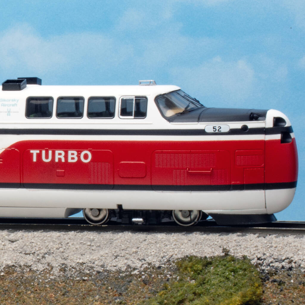 Futuristic toy train painted in red, white, and black colors on a model base with green grass and a tan concrete culvert in front of a blue sky backdrop with white fluffy clouds.