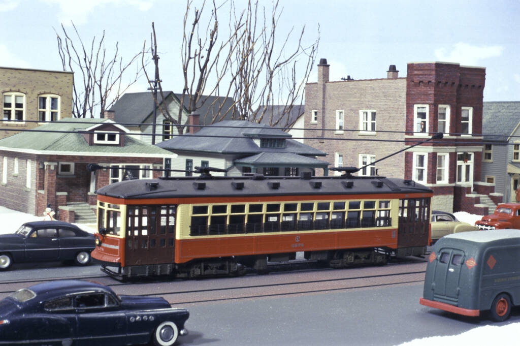 A street car passes houses and cars in a neighborhood on a model railroad layout