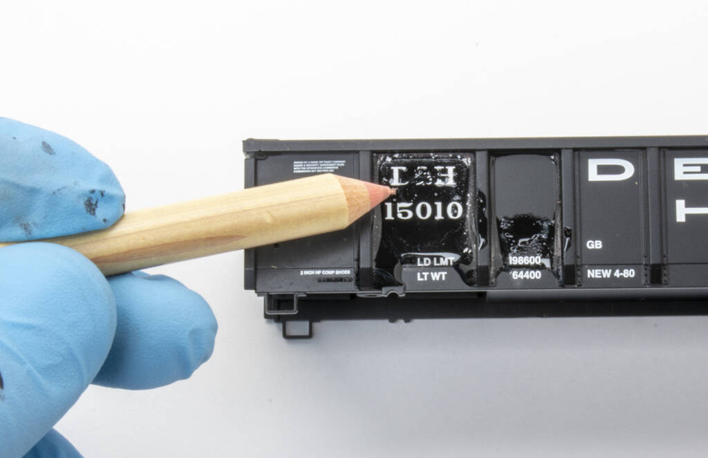 Color photo of black HO scale gondola with white lettering and gloved hand holding eraser pencil removing factory lettering