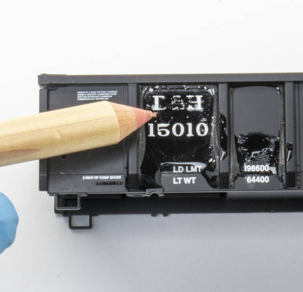 Color photo of black HO scale gondola with white lettering and gloved hand holding eraser pencil removing factory lettering